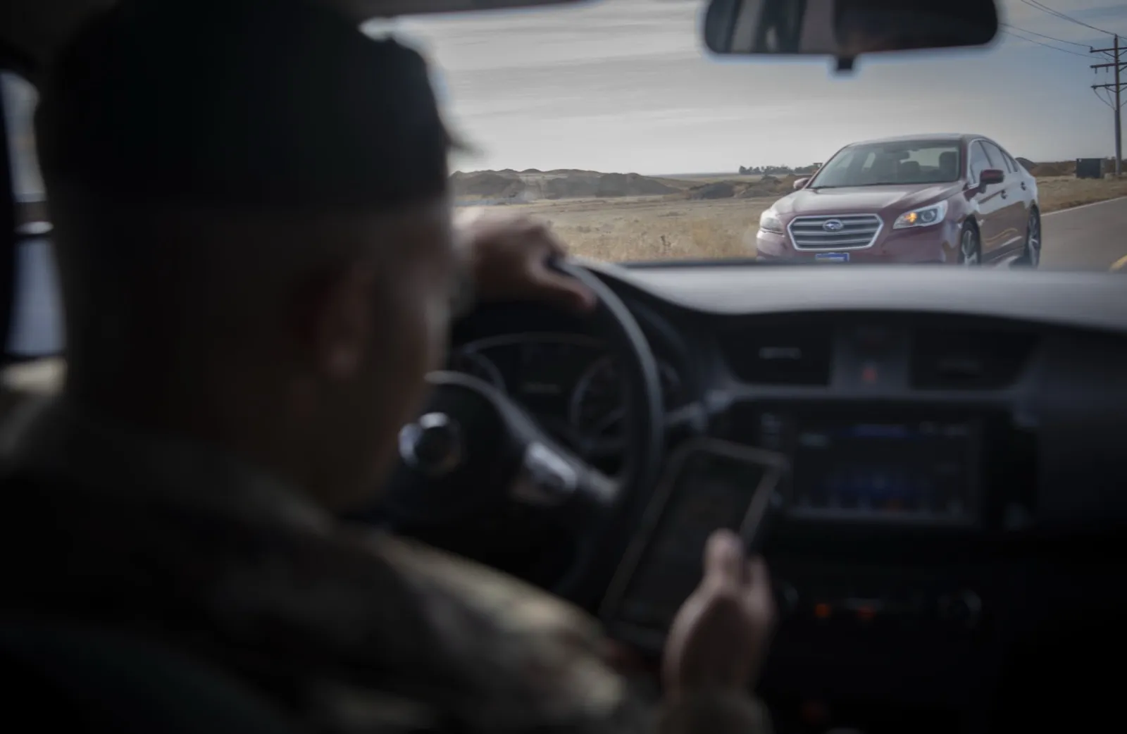 Seen from behind, a driver holds a phone low beside the steering wheel, eyes down, while a red car comes toward them on the road ahead